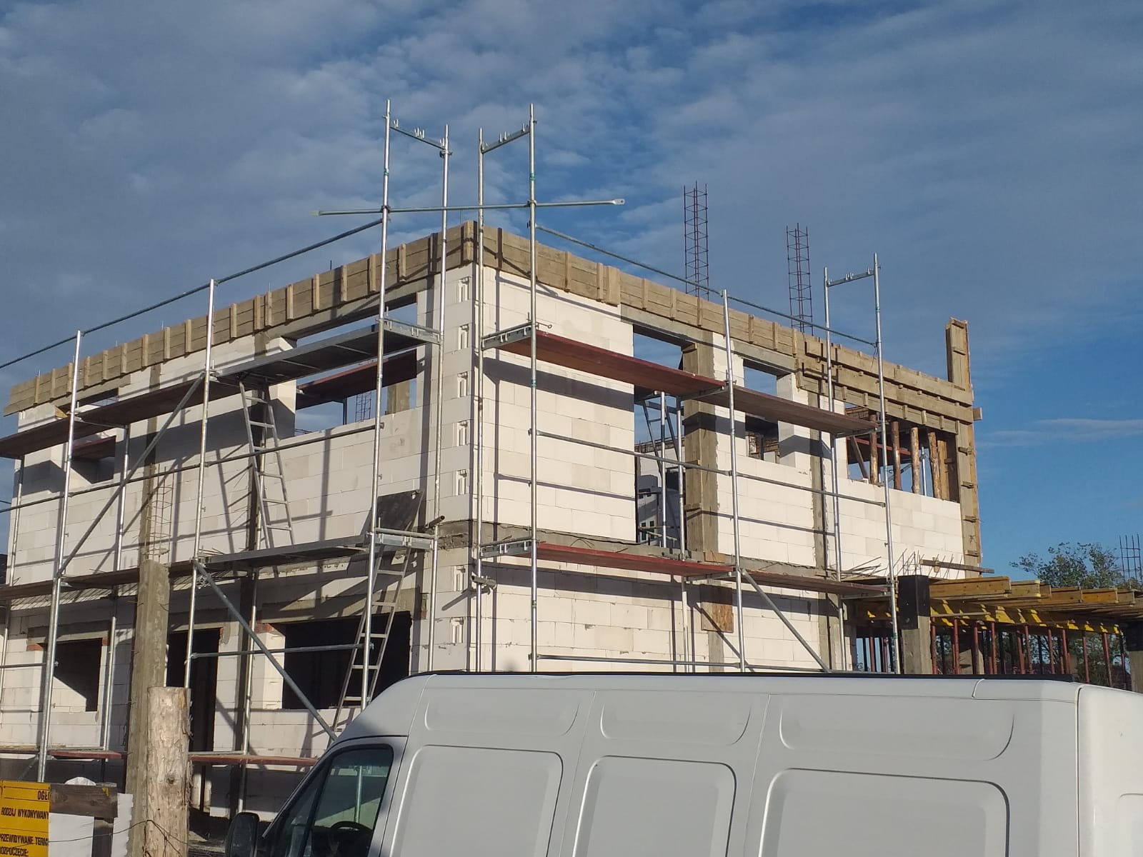 Wznoszenie dwukondygnacyjnego budynku z białych bloczków, widoczne rusztowania, szalunki na stropie oraz zaparkowany biały bus.
