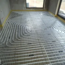 Rozłożone rury ogrzewania podłogowego na metalowej siatce w surowym pomieszczeniu z oknami, przygotowane do zalania wylewką.