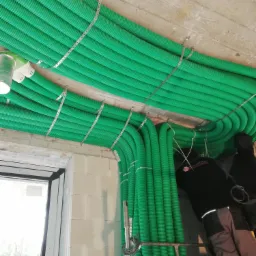 Zielone, karbowane rury wentylacyjne zamontowane pod sufitem, przymocowane metalowymi taśmami perforowanymi, widoczny fragment okna i pracownik na drabinie w tle.