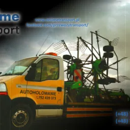 Żółto-biały bus z napisem 'AUTOHOLOWANIE' przewożący zielony sprzęt rolniczy na platformie, widok z boku na tle pochmurnego nieba.