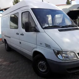 Biały furgon Mercedes-Benz Sprinter zaparkowany na brukowanym podjeździe, widok z przodu i boku, z widoczną tablicą rejestracyjną DGL 7...