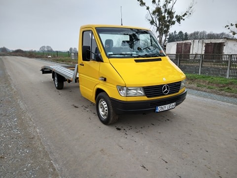 Żółty samochód ciężarowy Mercedes-Benz z platformą lawety, stojący na szutrowej drodze, widok z przodu, tablica rejestracyjna widoczna.