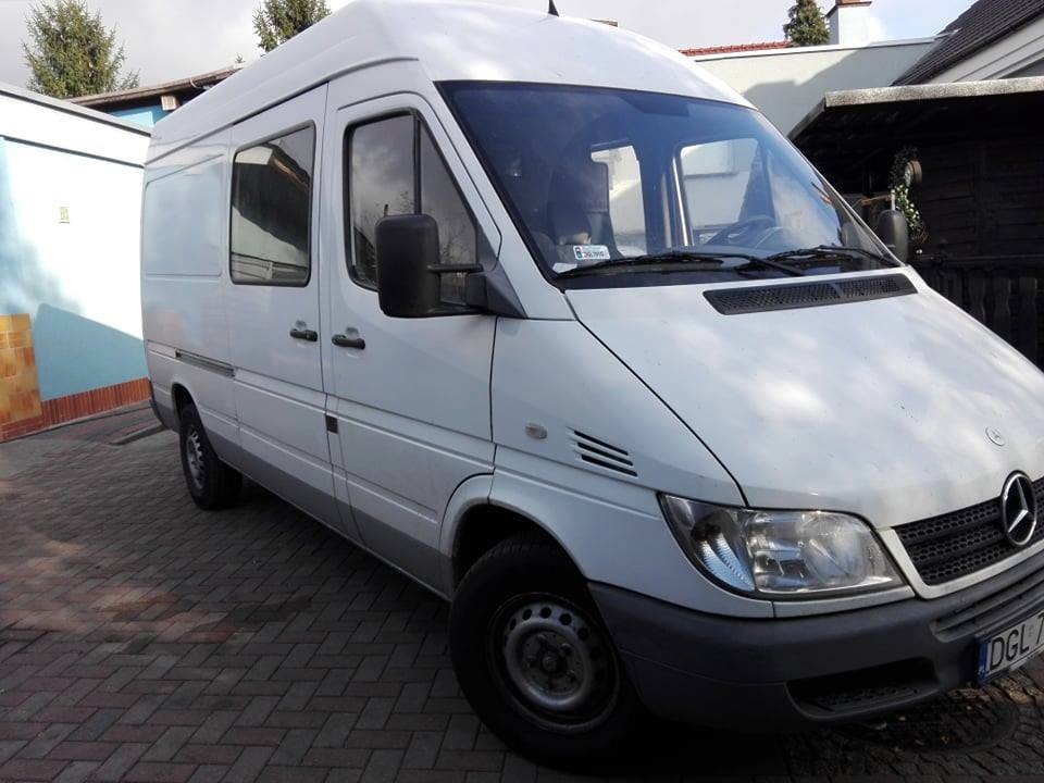 Biały furgon Mercedes-Benz Sprinter zaparkowany na brukowanym podjeździe, widok z przodu i boku, z widoczną tablicą rejestracyjną DGL 7...