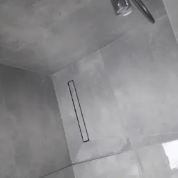 Szare płytki podłogowe i odpływ liniowy w kabinie prysznicowej w Pelplinie, widok z góry, chromowana słuchawka prysznicowa w tle.