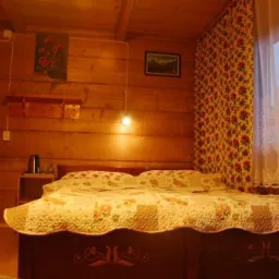 Drewniany pokój z podwójnym łóżkiem, patchworkową narzutą w kwiaty, wzorzystą tapetą i oknem z białymi, koronkowymi firankami.