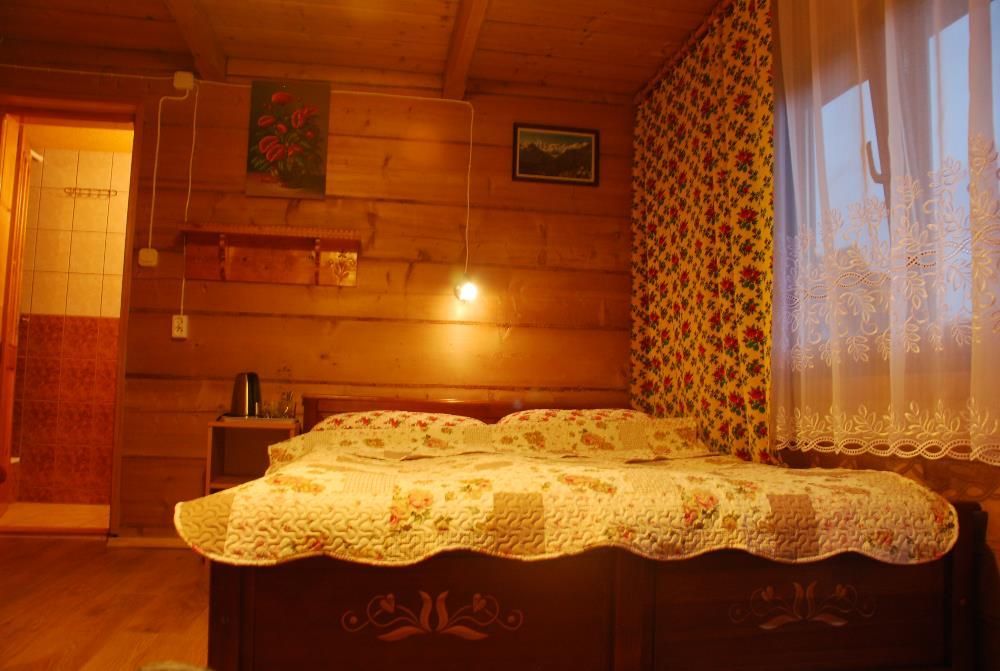 Drewniany pokój z podwójnym łóżkiem, patchworkową narzutą w kwiaty, wzorzystą tapetą i oknem z białymi, koronkowymi firankami.