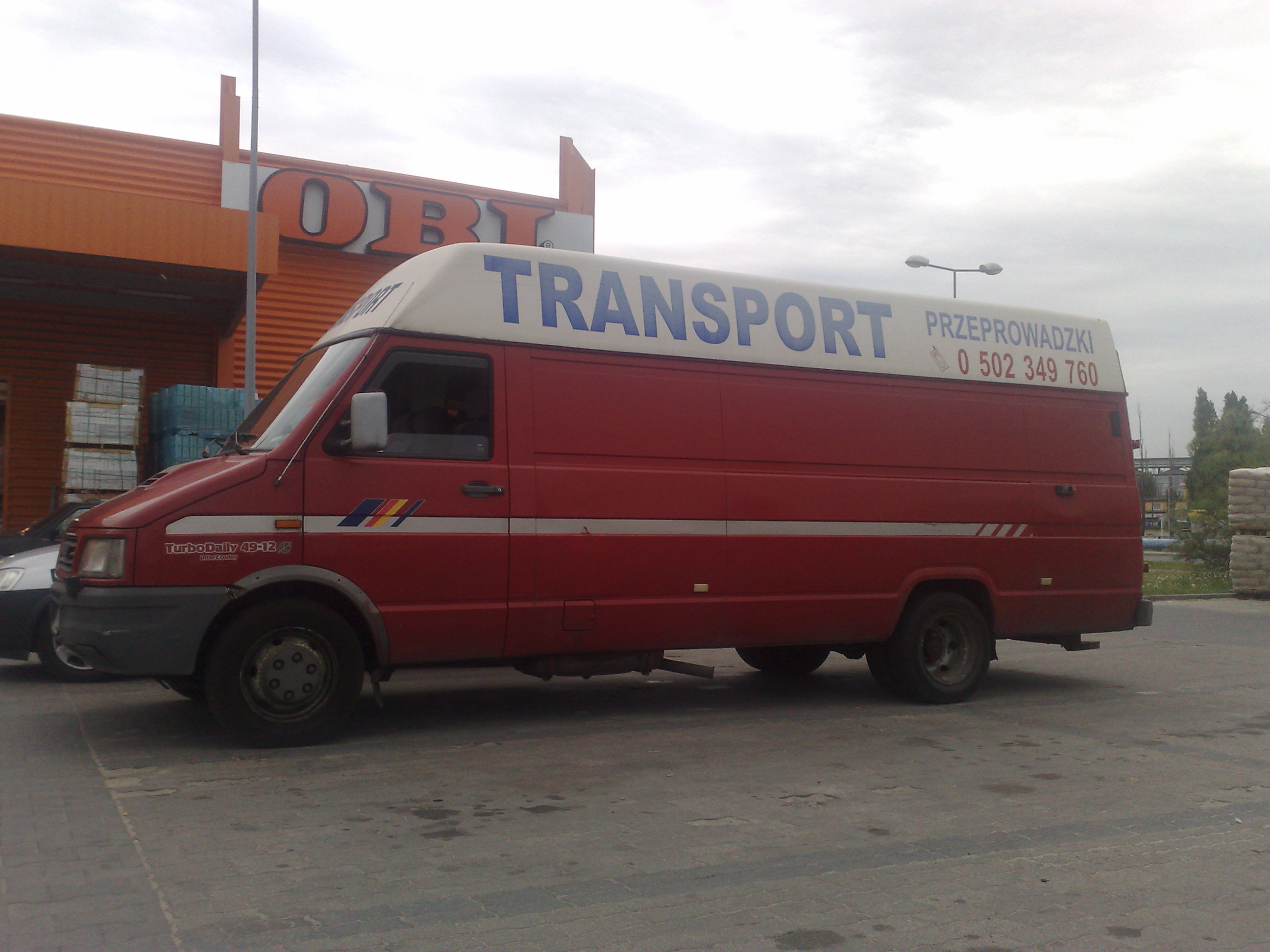 Czerwony samochód dostawczy z białym dachem, na którym widnieje napis 'TRANSPORT' i numer telefonu, zaparkowany na tle sklepu OBI.