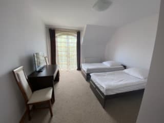 Pokój z dwoma pojedynczymi łóżkami, biurkiem z telewizorem i krzesłem. Widok z perspektywy wejścia, stonowane kolory, zasłonięte okno.