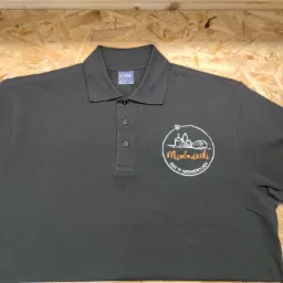 Czarna koszulka polo marki Uneek z nadrukiem logo 'Miodowianki KGW w Miedniewicach' w kolorze białym i pomarańczowym, leżąca na drewnianym tle.