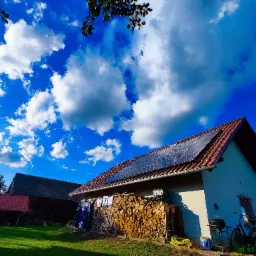 Dom jednorodzinny z panelami słonecznymi na dachu, stosem drewna opałowego i suszącym się praniem w słoneczny dzień z intensywnie błękitnym niebem i białymi chmurami.