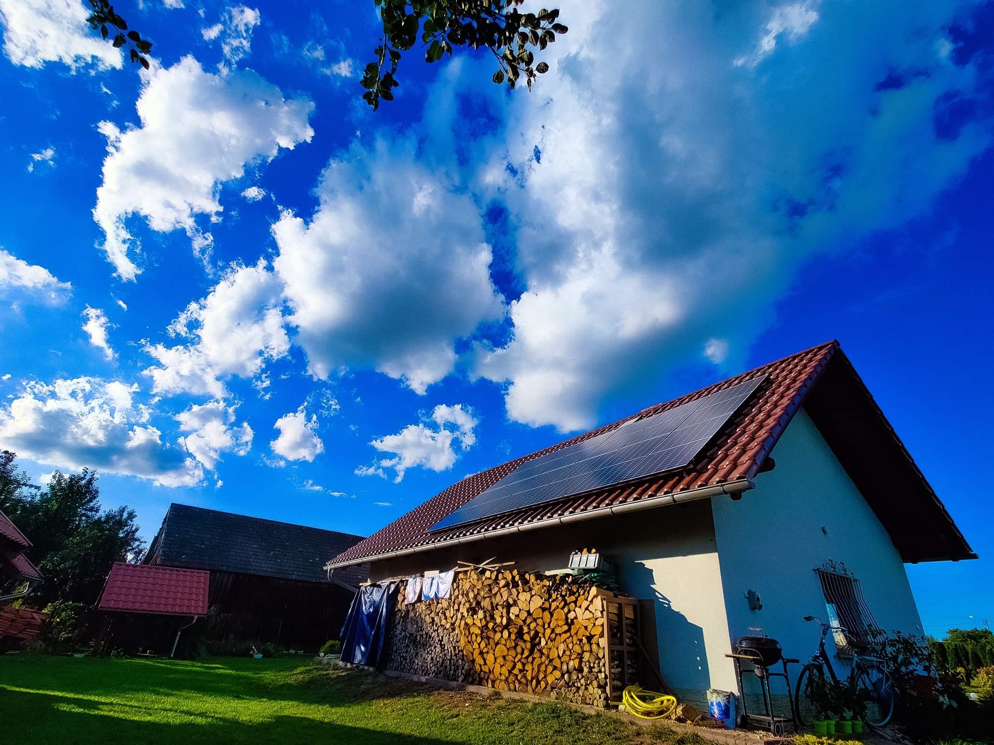 Dom jednorodzinny z panelami słonecznymi na dachu, stosem drewna opałowego i suszącym się praniem w słoneczny dzień z intensywnie błękitnym niebem i białymi chmurami.