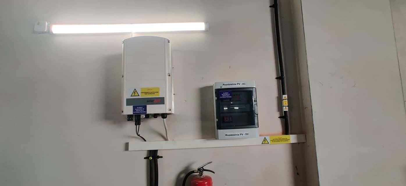 Biały inwerter fotowoltaiczny i rozdzielnica PV AC/DC zamontowane na ścianie w pomieszczeniu technicznym, widoczny gaśnica w dolnym rogu.