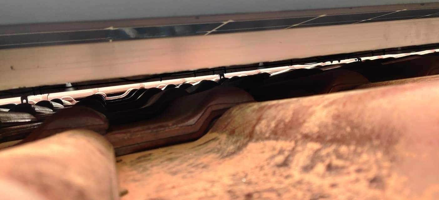 Widok spod paneli słonecznych zamontowanych na dachu z czerwonej dachówki, widoczne przewody i elementy konstrukcyjne mocujące panele.