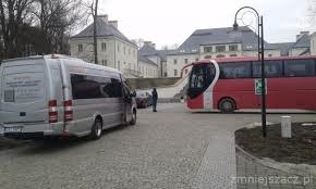 Dwa autokary (srebrny bus i czerwony autobus) stojące na brukowanym placu przed dużym, jasnym budynkiem z wieloma oknami. Widoczna osoba stojąca obok samochodu osobowego. Na srebrnym busie widoczne...