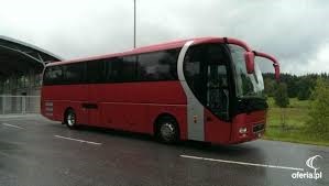 Czerwony autokar turystyczny MAN na mokrym asfalcie, widok z boku, zarys budynku w tle.