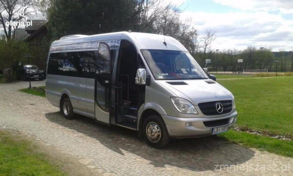 Srebrny Mercedes Sprinter z otwartymi drzwiami pasażera, zaparkowany na żwirowej nawierzchni w otoczeniu zieleni, z widocznym napisem 'WYNAJEM 2504 245' na szybie.