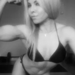 Czarno-białe zdjęcie kobiety w stroju sportowym, prezentującej napięte bicepsy na tle domowej siłowni z obciążeniami.