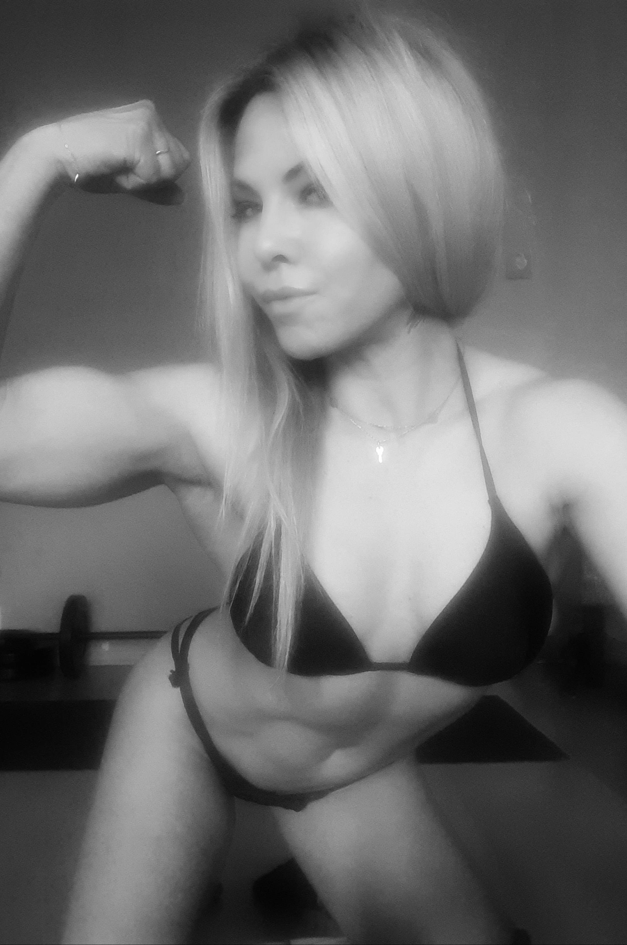 Czarno-białe zdjęcie kobiety w stroju sportowym, prezentującej napięte bicepsy na tle domowej siłowni z obciążeniami.