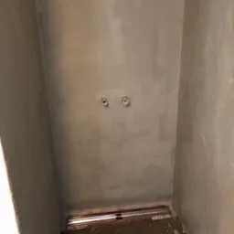 Przygotowana do prac wykończeniowych, surowa ściana w łazience z wyprowadzonymi rurami instalacji wodnej, widoczna listwa przypodłogowa i fragment podłogi.