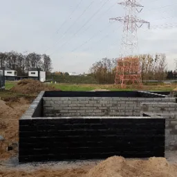 Wznoszenie fundamentów z bloczków betonowych, zabezpieczonych czarną izolacją, na tle krajobrazu z liniami wysokiego napięcia i nowoczesnymi domami.
