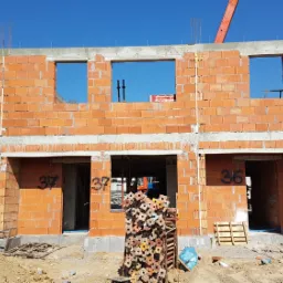 Wznoszenie ścian z czerwonej cegły Porotherm na budowie szeregowca, widoczne numery domów, stemple budowlane i żuraw budowlany w tle.