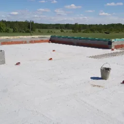 Fundamenty domu z betonu i cegły ceramicznej, widok z góry na plac budowy z wiadrami i miotłą, w tle zielone drzewa i błękitne niebo.