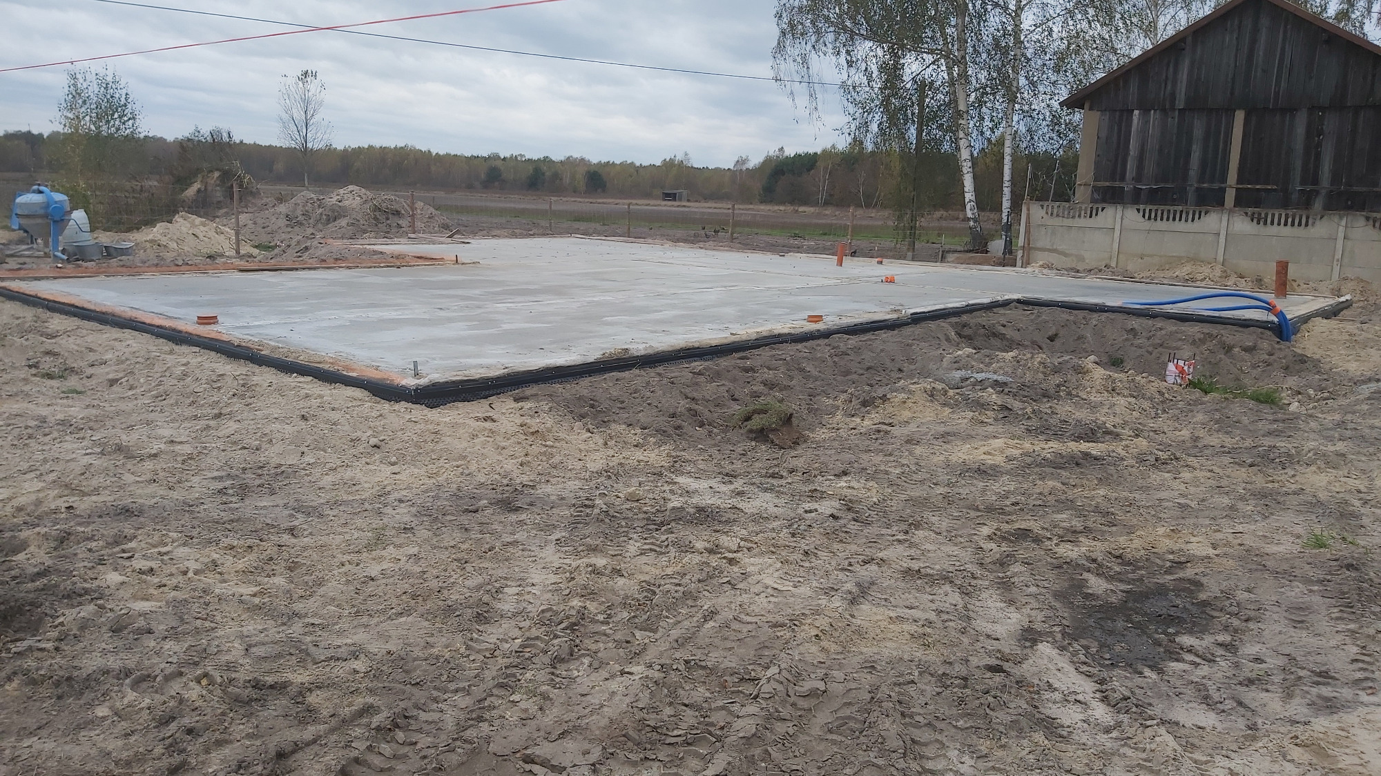 Wylany betonowy fundament pod budynek, widoczne rury instalacyjne i czarna izolacja pionowa, w tle mieszalnik betonu i stary budynek gospodarczy.