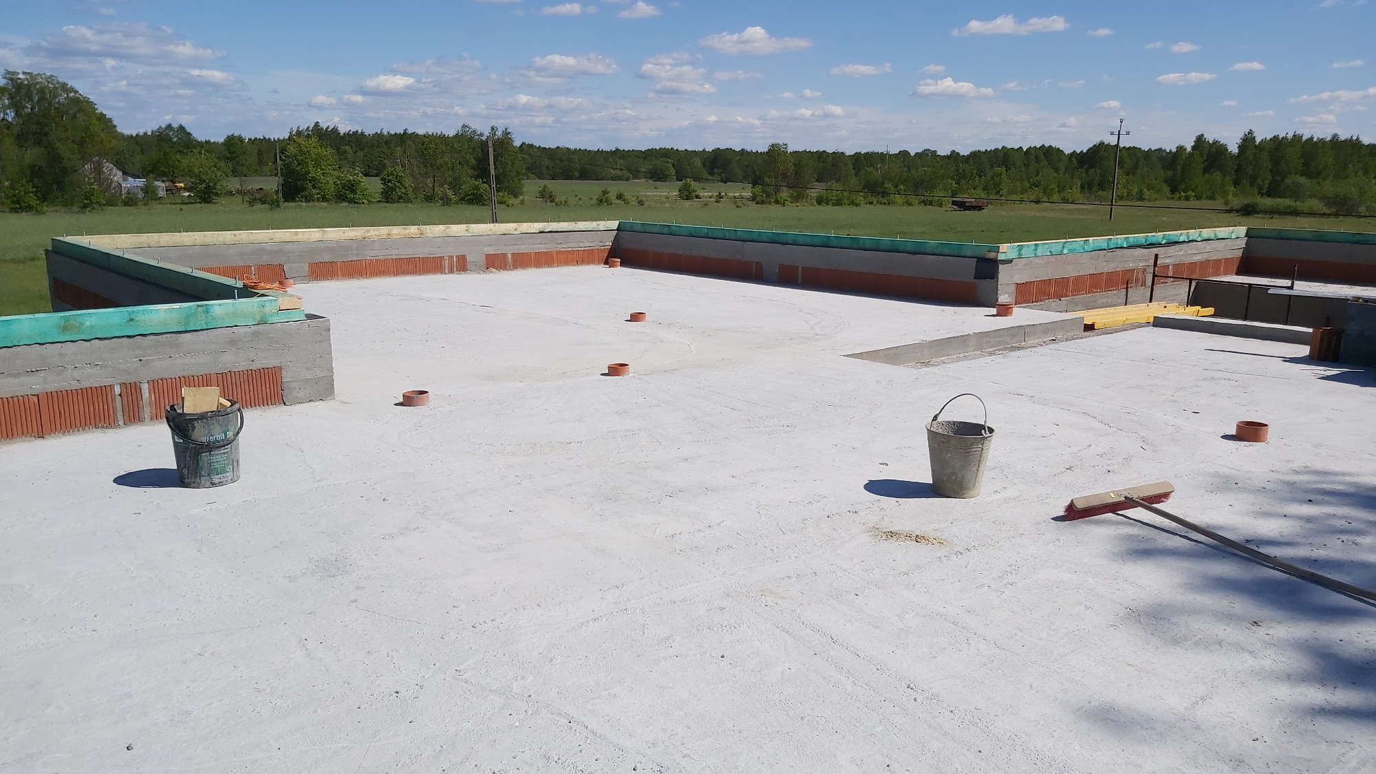 Fundamenty domu z betonu i cegły ceramicznej, widok z góry na plac budowy z wiadrami i miotłą, w tle zielone drzewa i błękitne niebo.