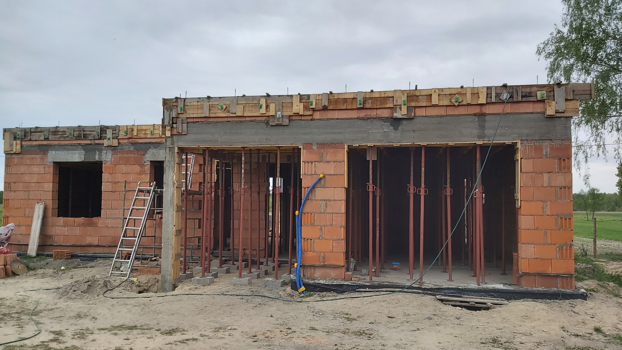 Budowa domu murowanego w stanie surowym otwartym: widoczne ściany z czerwonej cegły, drewniane szalunki, stalowe podpory i drabina aluminiowa na tle pochmurnego nieba i zielonego krajobrazu.