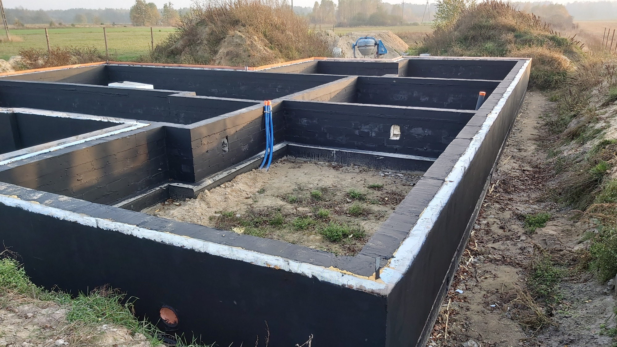 Zalane fundamenty pod budynek, zabezpieczone czarną izolacją, z widocznymi niebieskimi rurami instalacji wodnej i wylewką wyrównującą.