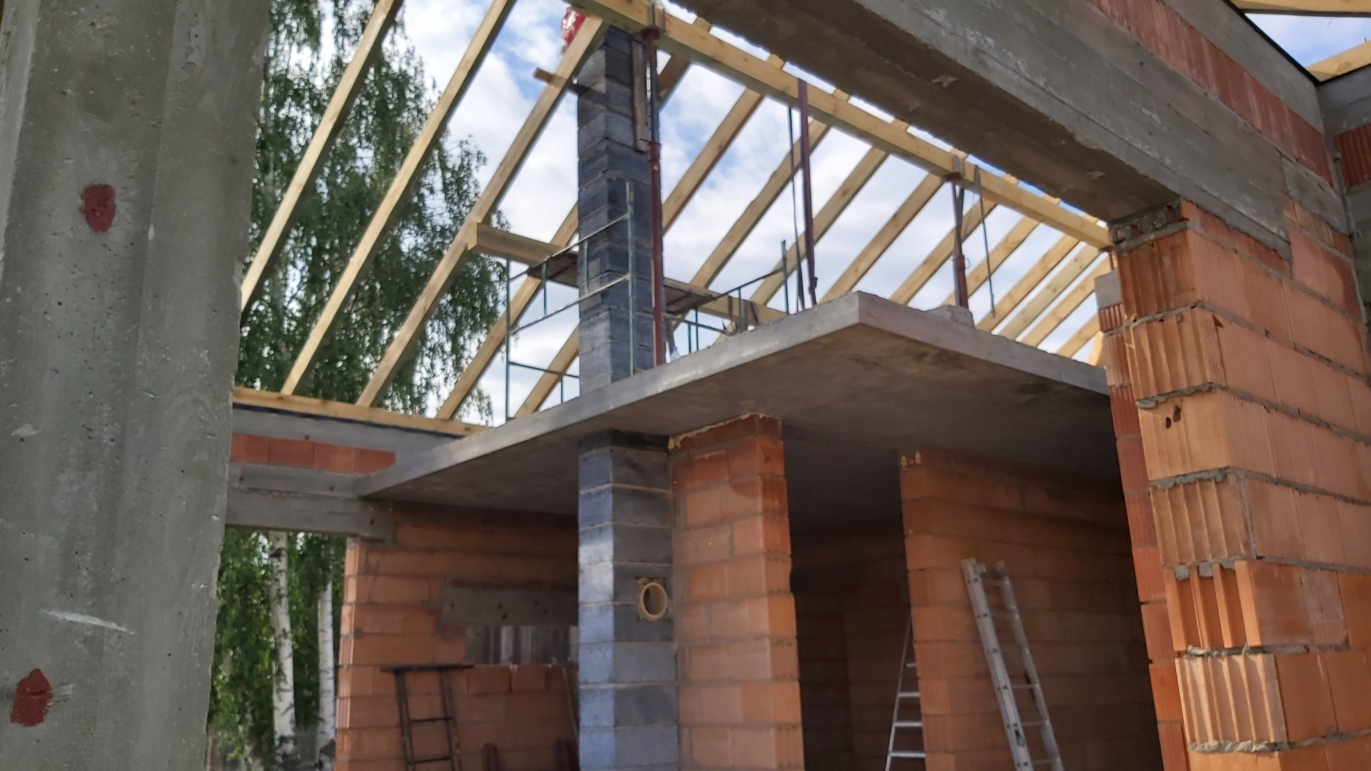 Surowy stan domu w trakcie budowy: widoczne ściany z czerwonej cegły, betonowe stropy, komin z szarych bloczków oraz drewniana konstrukcja dachu na tle błękitnego nieba.