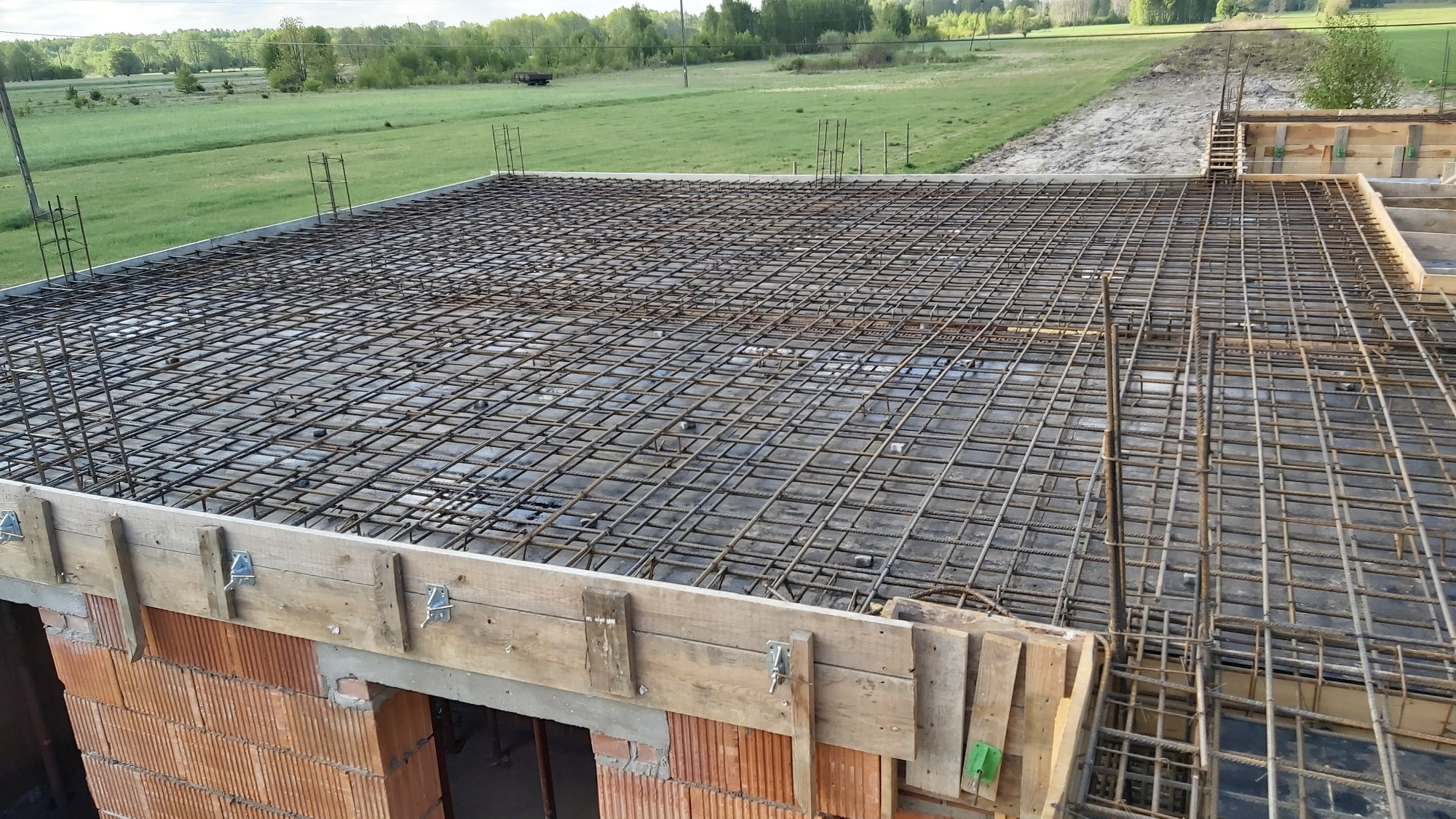Zbrojenie stropu domu z czerwonej cegły, widoczne drewniane szalunki i stalowe pręty tworzące siatkę zbrojeniową na tle zielonego pola.