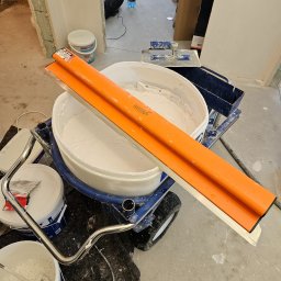 Marcin Owczarek - roboty ogólnobudowlane - Przygotowanie do nakładania gładzi w Szczecinie: wiadro z masą szpachlową na wózku, obok narzędzia do nakładania, widoczne ślady prac remontowych.