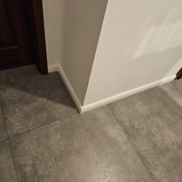 Marcin Owczarek - roboty ogólnobudowlane - Świeżo pomalowana ściana w rogu pomieszczenia z listwą przypodłogową w kolorze białym, przylegająca do ciemnobrązowej futryny drzwiowej, na podłodze szare płytki imitujące beton.