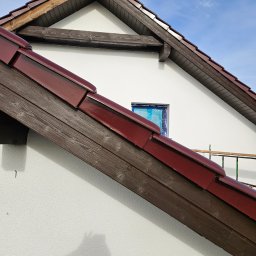 Marcin Owczarek - roboty ogólnobudowlane - Fragment elewacji domu z widocznym oknem zabezpieczonym folią malarską, świeżo pomalowaną białą ścianą i brązową dachówką. Na ścianie widoczny cień osoby robiącej zdjęcie.