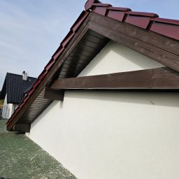Marcin Owczarek - roboty ogólnobudowlane - Wykończona elewacja domu z widocznym fragmentem dachu pokrytego czerwoną dachówką, brązowe elementy drewniane i biała, świeżo nałożona struktura tynku na ścianie. Widoczny fragment dachu...