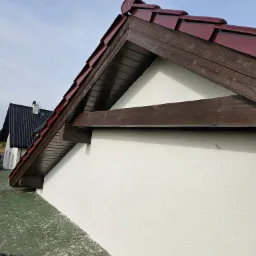 Wykończona elewacja domu z widocznym fragmentem dachu pokrytego czerwoną dach&oacute;wką, brązowe elementy drewniane i biała, świeżo nałożona struktura tynku na ścianie. Widoczny fragment dachu...