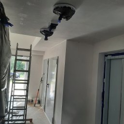 Marcin Owczarek - roboty ogólnobudowlane - Korytarz w trakcie malowania: zabezpieczone lampy, drabina oparta o ścianę, folia ochronna na podłodze, świeżo pomalowane ściany i sufit.