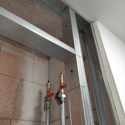 Marcin Owczarek - roboty ogólnobudowlane - Montaż metalowej konstrukcji pod płytę karton gips na nierównej ścianie z widocznymi rurami instalacji wodnej zakończonymi zaworami.