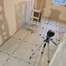 Układanie płytek podłogowych w pomieszczeniu, widoczny laser krzyżowy na statywie, drabina aluminiowa i fragment ściany po szpachlowaniu.