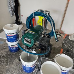 Marcin Owczarek - roboty ogólnobudowlane - Agregat malarski Grone PowerSpray z niebieskim wężem i kilkoma wiadrami z gładzią szpachlową Master Mas Classic na folii ochronnej w trakcie prac wykończeniowych.