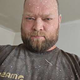 Marcin Owczarek - roboty ogólnobudowlane - Mężczyzna z brodą, pokryty białymi plamami farby lub gładzi, spogląda z góry, ubrany w brudną koszulkę z logo firmy budowlanej.