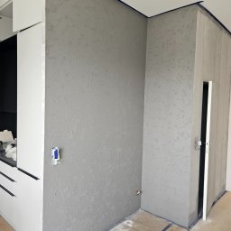 Marcin Owczarek - roboty ogólnobudowlane - Świeżo położona, szara struktura na ścianie w rogu pomieszczenia, zabezpieczona niebieską taśmą malarską przy suficie i podłodze, z widoczną białą zabudową kuchenną po lewej stronie i otwartymi...