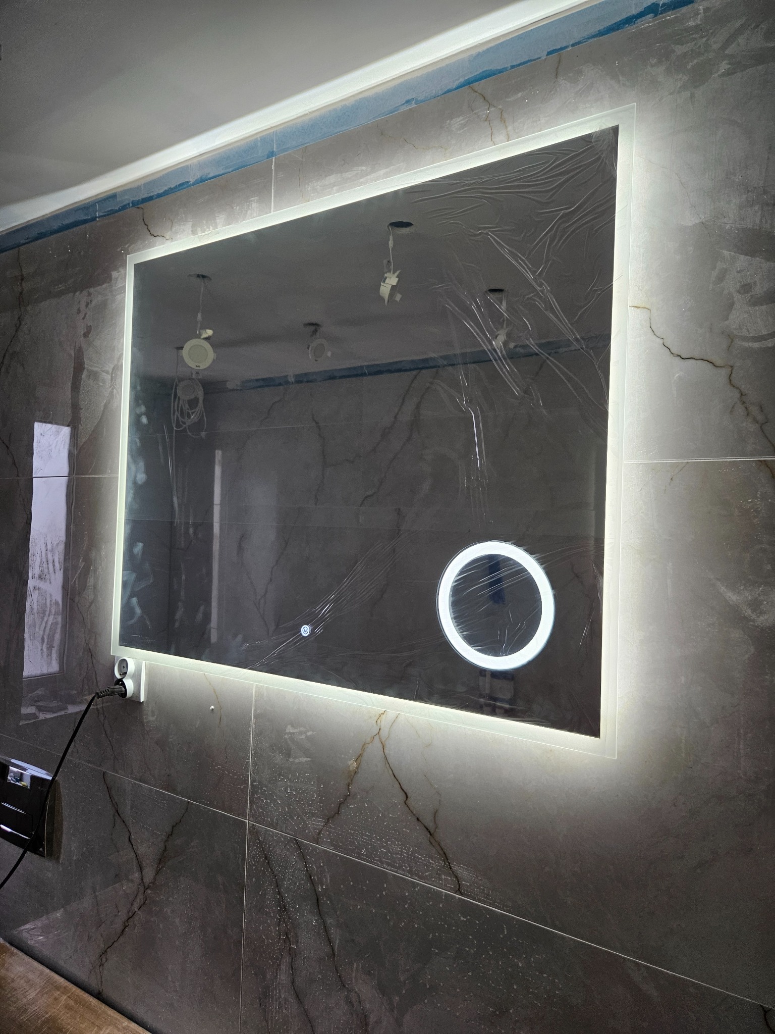 Podświetlane lustro prostokątne z okrągłym dodatkowym oświetleniem, zamontowane na ścianie wyłożonej szarymi płytkami imitującymi marmur, zabezpieczone folią ochronną, widoczne kable i otwory...