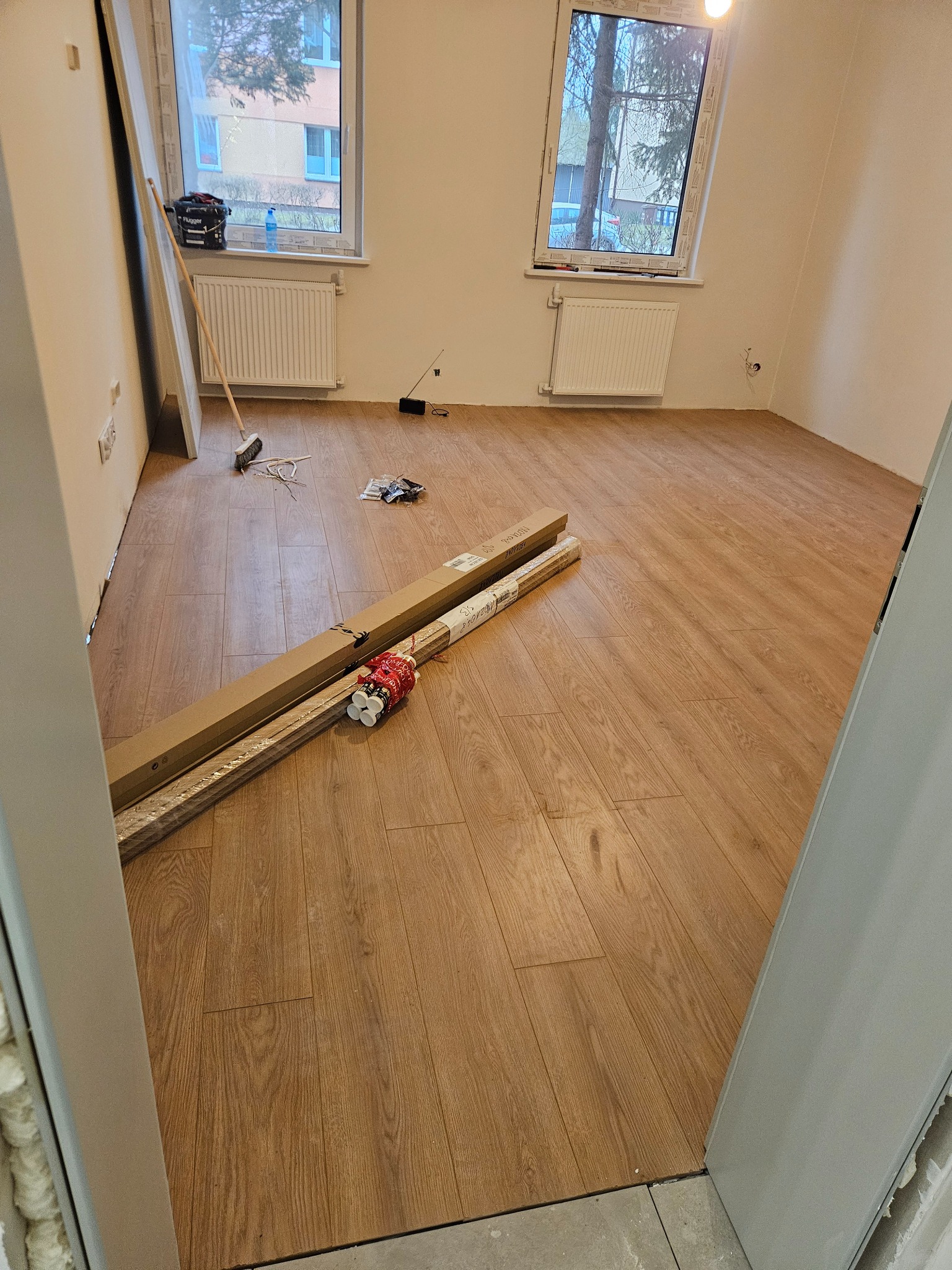 Nowa podłoga laminowana w pokoju podczas układania, widoczne paczki paneli i listew przypodłogowych, szczotka, okna z grzejnikami i częściowo wykończone ściany.