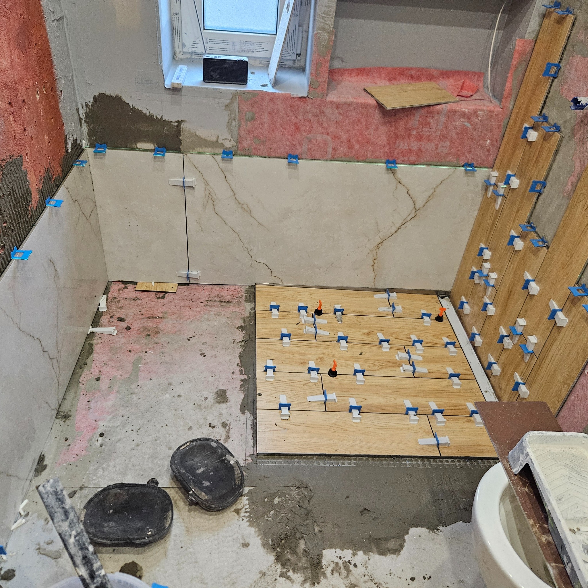 Łazienka w trakcie układania płytek imitujących drewno na podłodze i marmur na ścianach, widoczne narzędzia, poziomice i system poziomowania płytek.