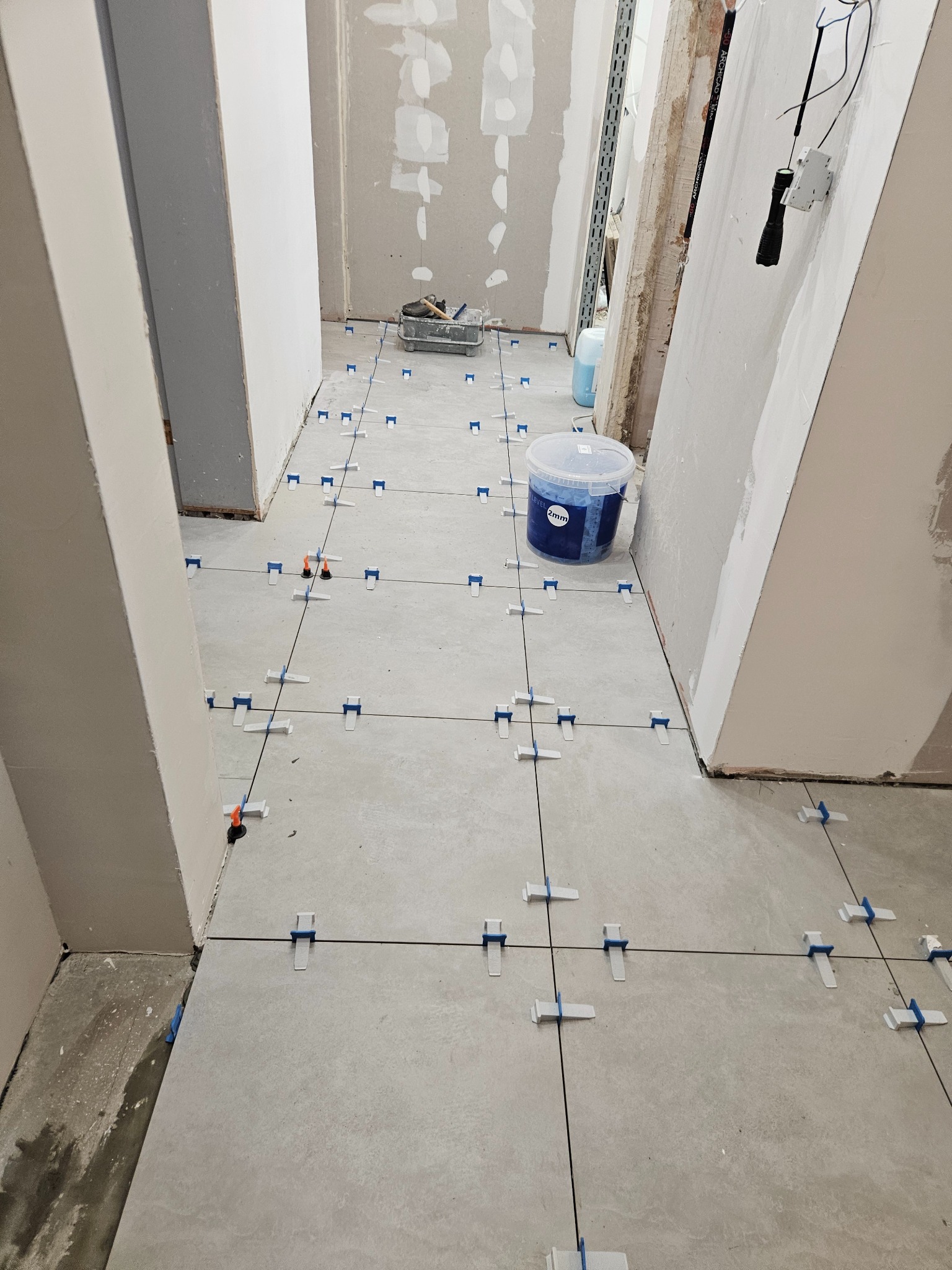 Układanie szarych płytek podłogowych z systemem poziomowania w korytarzu w Szczecinie, widoczny pojemnik z klejem i narzędzia.