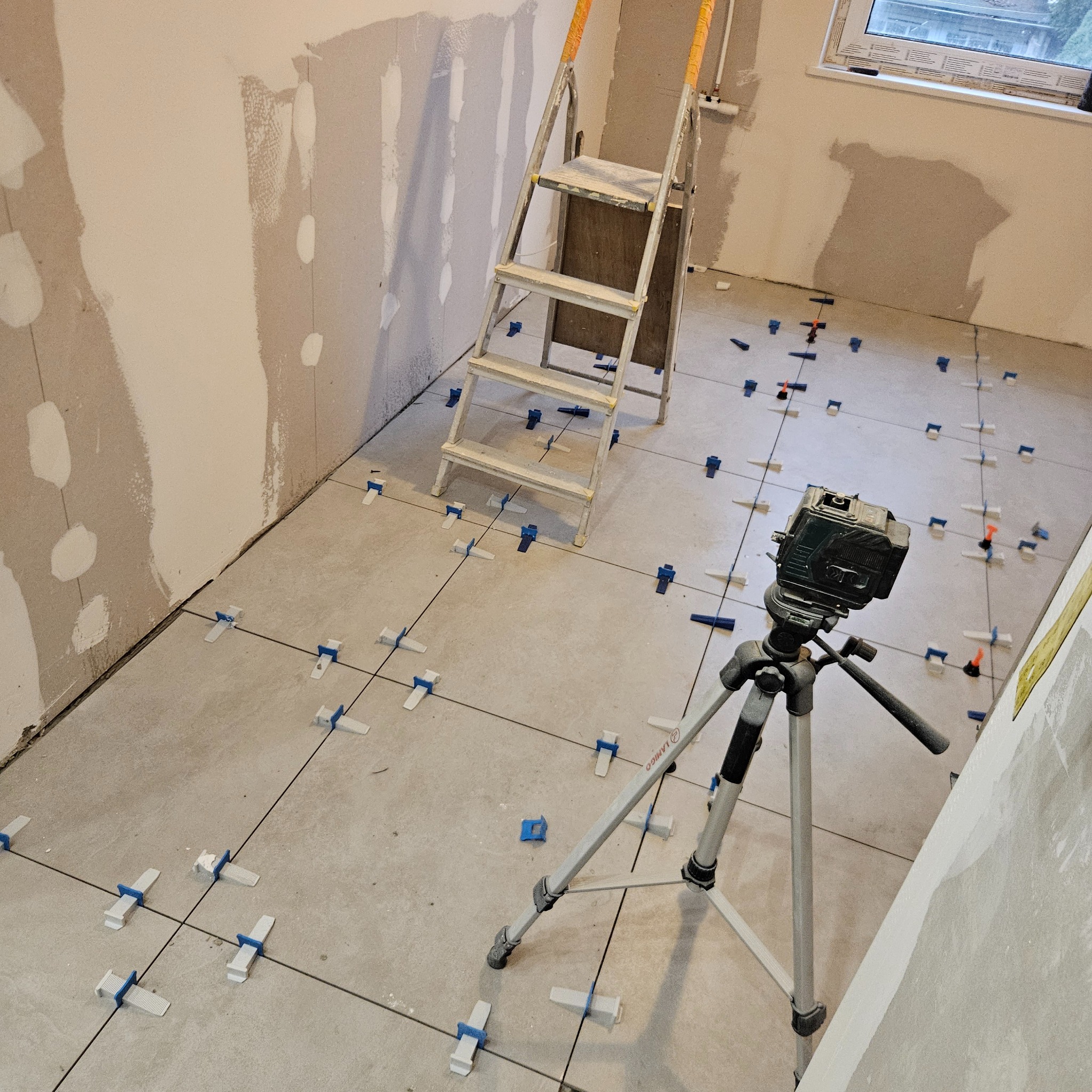 Układanie płytek podłogowych w pomieszczeniu, widoczny laser krzyżowy na statywie, drabina aluminiowa i fragment ściany po szpachlowaniu.