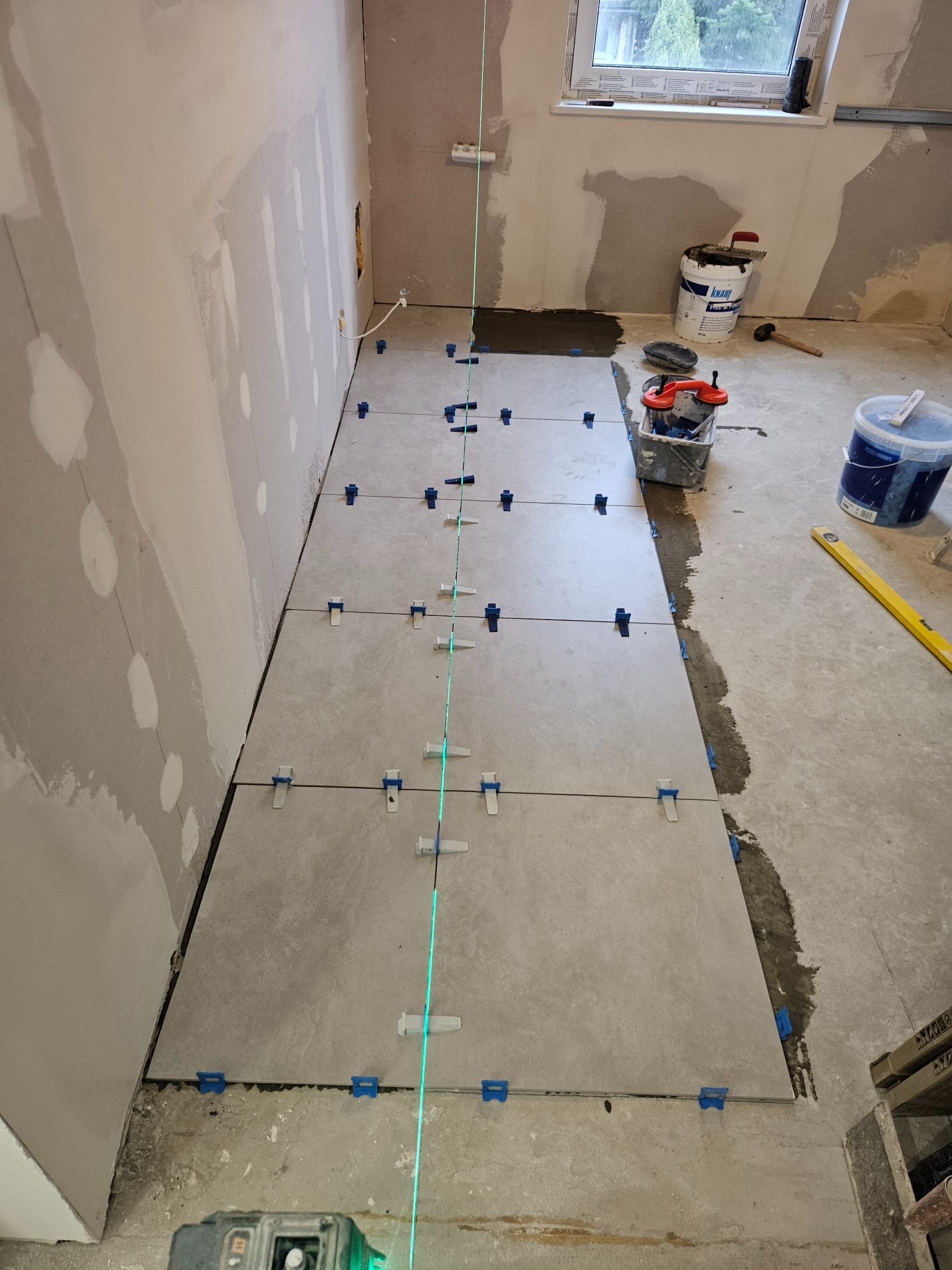 Układanie szarych płytek podłogowych z użyciem poziomicy laserowej i klipsów poziomujących, ściany przygotowane do malowania, widoczne narzędzia i pojemniki z zaprawą.