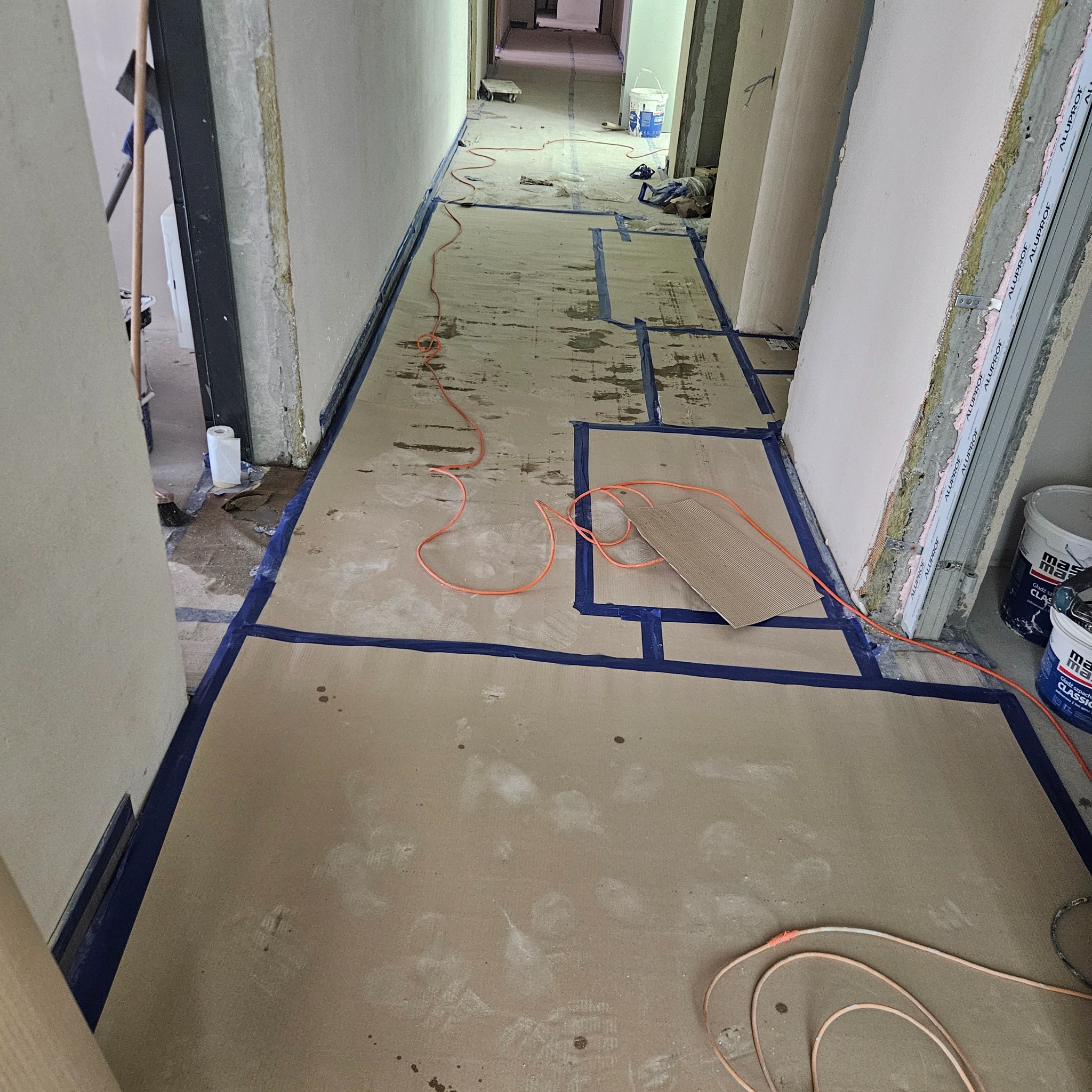 Korytarz w trakcie przygotowania do prac wykończeniowych: podłoga zabezpieczona kartonem i taśmą malarską, ściany częściowo z płyt kartonowo-gipsowych, widoczne wiadra z masą szpachlową i przewody...